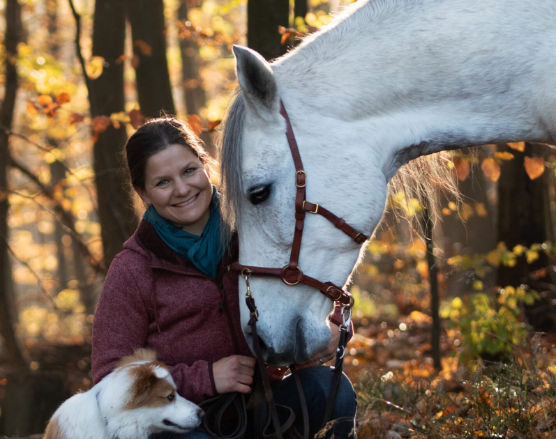 Tierärztin und Chiropraktikerin Silke Paret im vertrauten Kontakt mit einem weißen Pferd und ihrem Hund im herbstlichen Wald